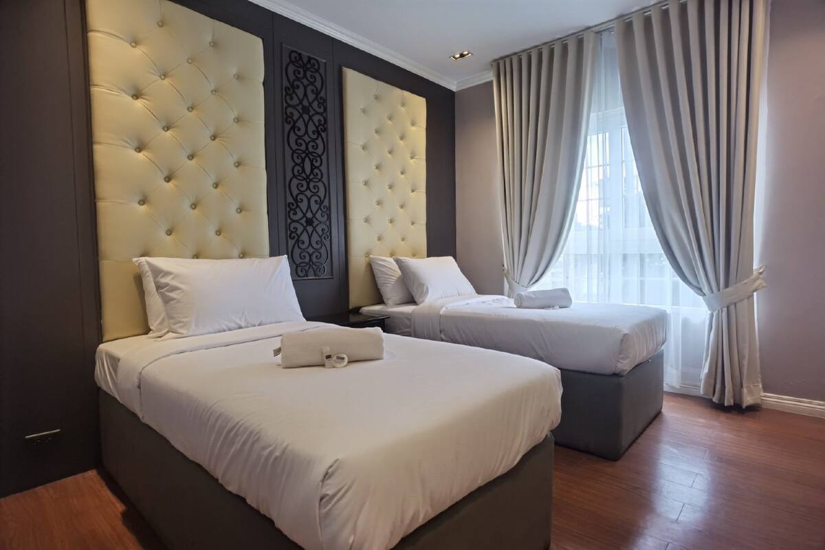 A hotel room with two beds, a large tufted headboard, a decorative wall panel, and a large window with curtains.