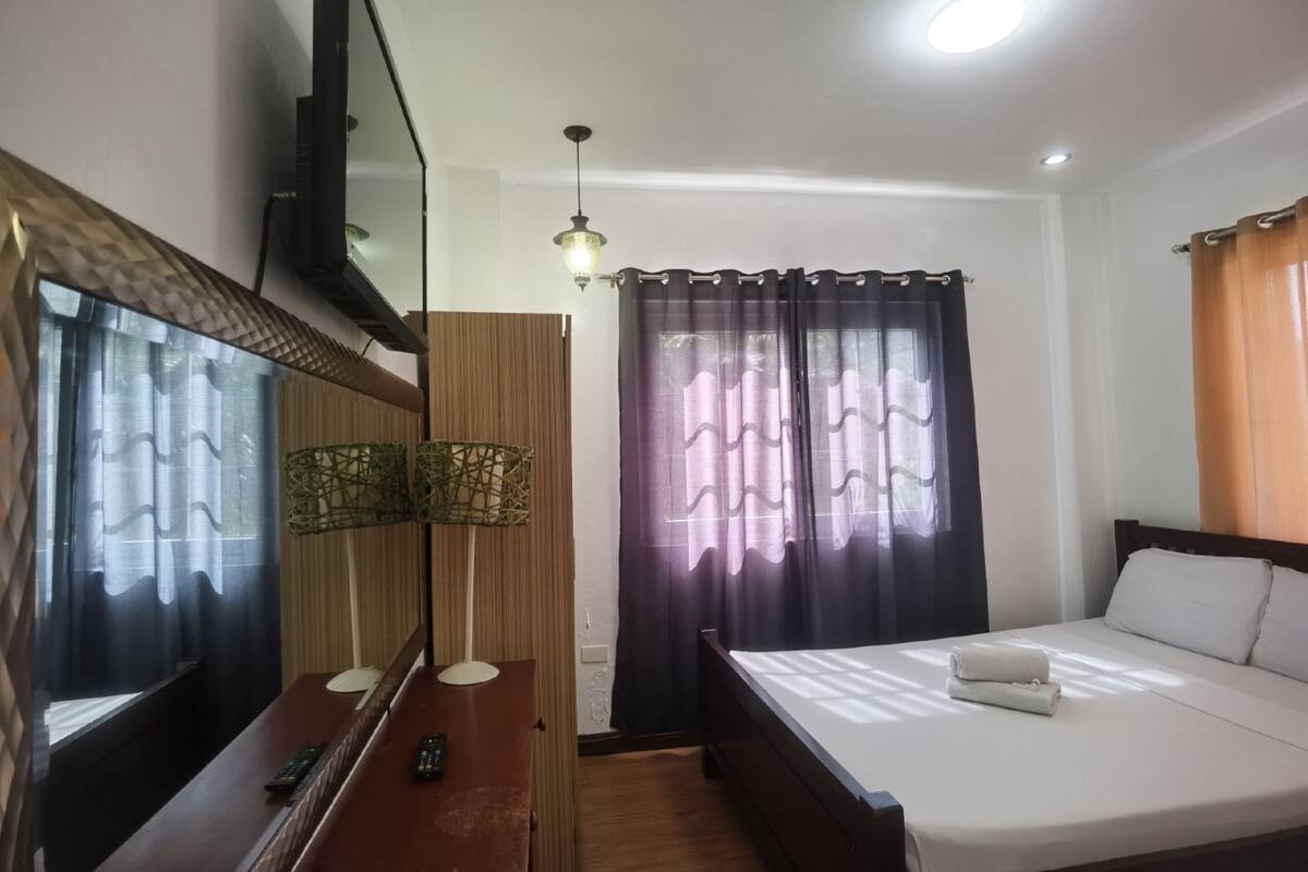 A hotel room with a bed, a desk with a lamp, a TV mounted on the wall, and a window with curtains.