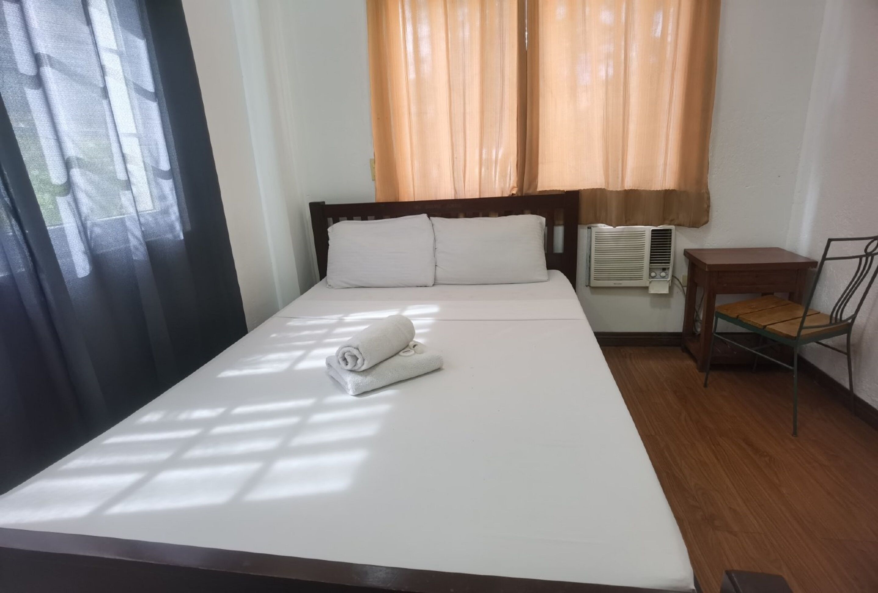 Una cama bien hecha con sábanas blancas, cabecera de madera y una toalla enrollada sobre la cama. Junto a la cama hay una pequeña mesa de madera con una silla, y un equipo de aire acondicionado en la pared.
