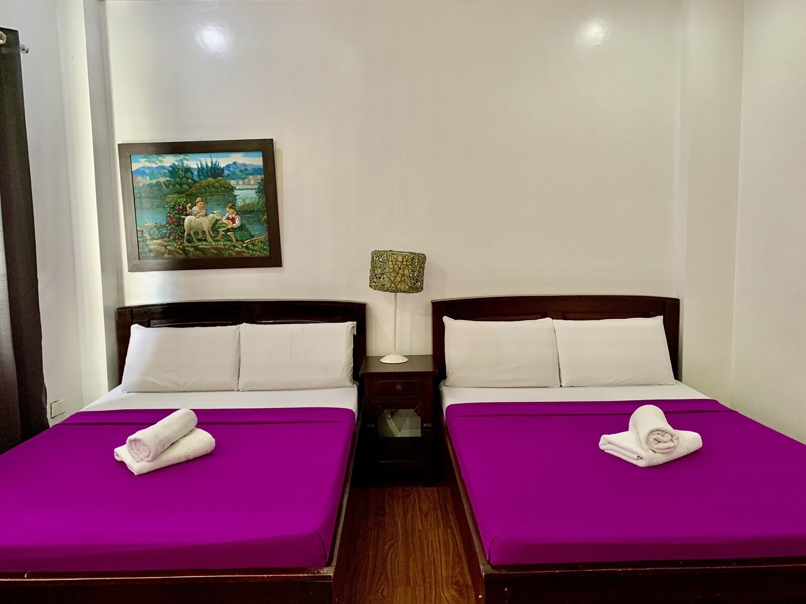 A hotel room with two beds, each with a purple bedspread and white pillows, a nightstand with a lamp, and a painting on the wall.