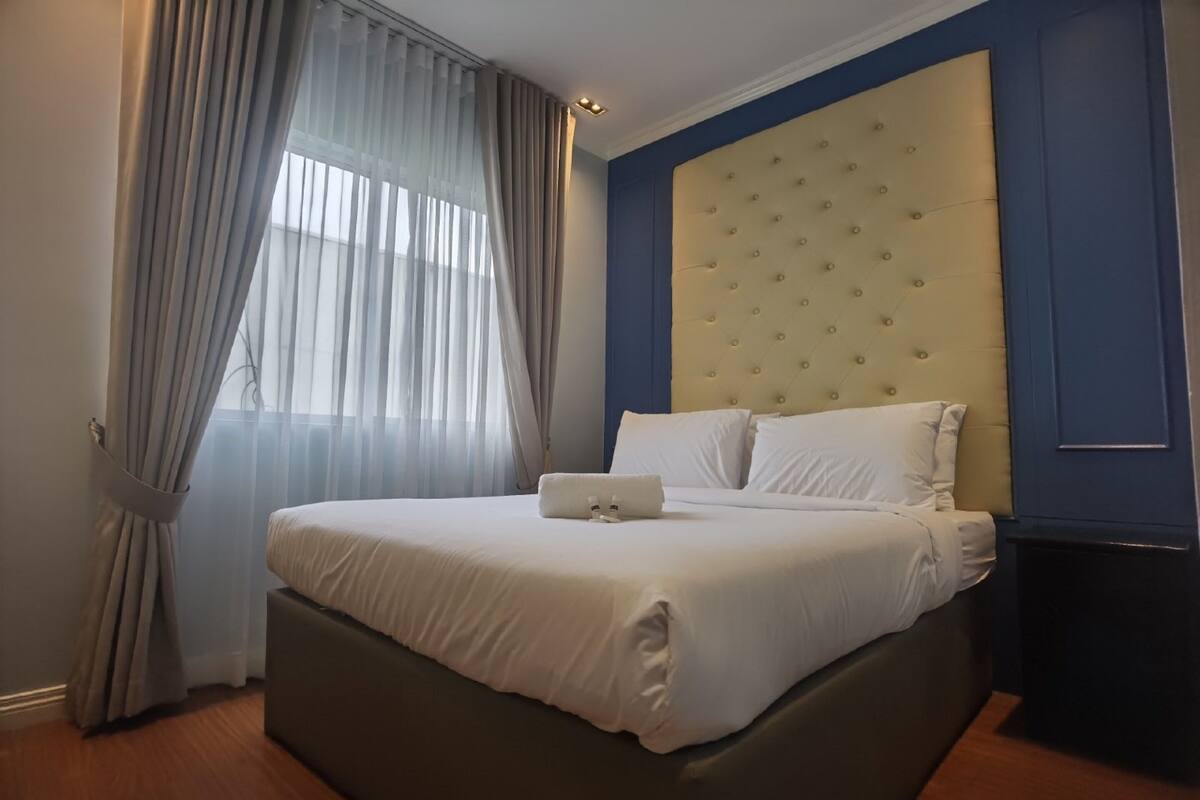 A neatly made bed with white linens and a white pillow, a tufted headboard, and a window with sheer curtains.