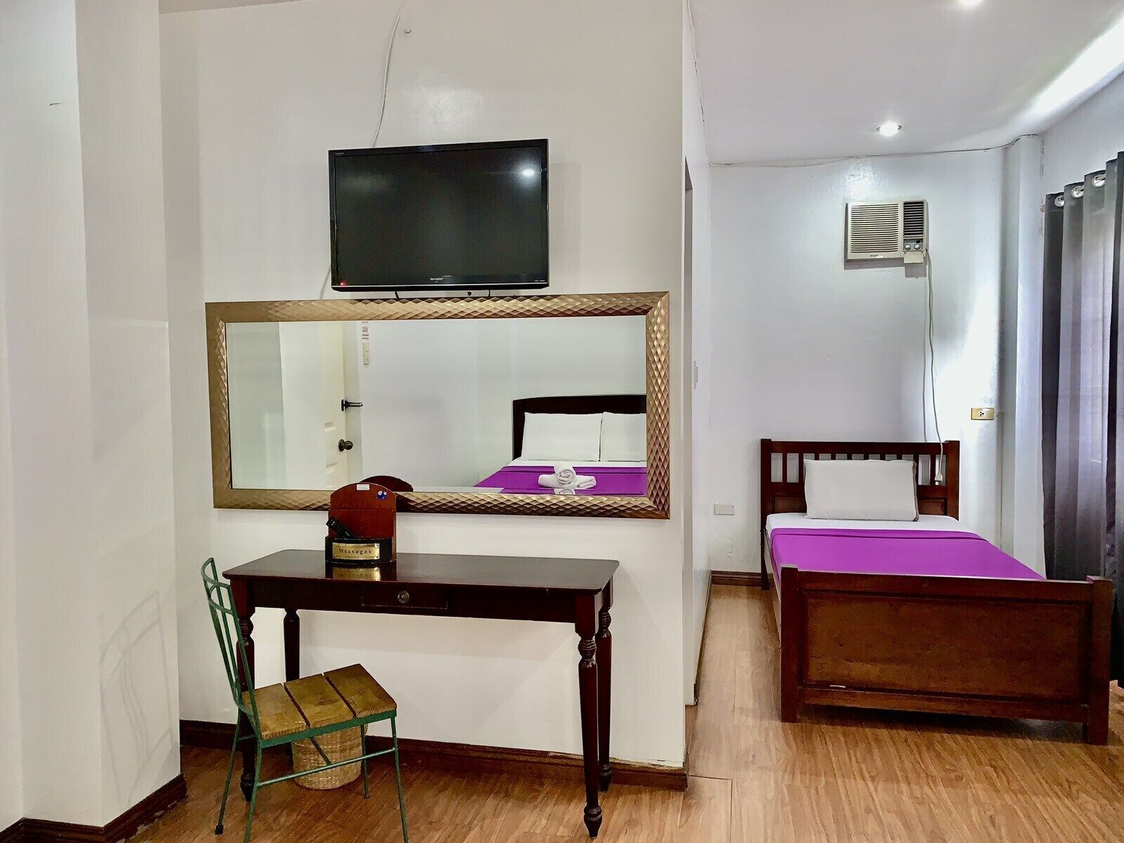 A hotel room with a wooden desk, a chair, a bed with a purple bedspread, a large mirror, a television, and an air conditioning unit.