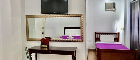 A hotel room with a wooden desk, a chair, a bed with a purple bedspread, a large mirror, a television, and an air conditioning unit.