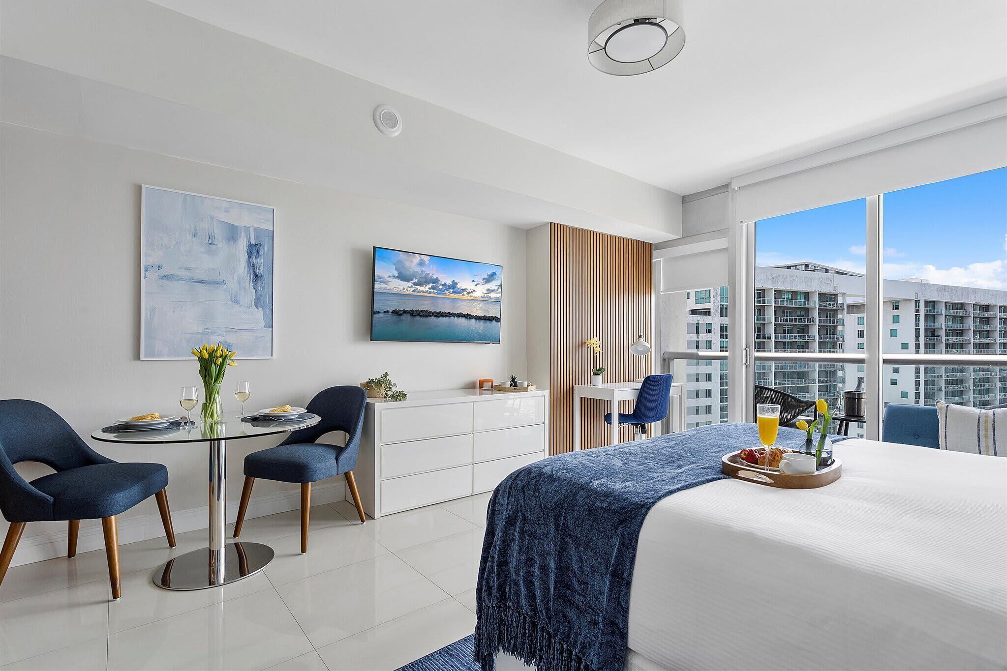 A modern hotel room with a large bed, a dining table with chairs, a desk, and a view of the cityscape.