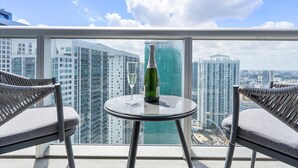 En balkon med to fletstole, et lille rundt bord, en champagneflaske og et glas. Udsigten inkluderer høje bygninger og et vandareal.