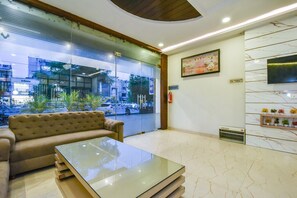 A hotel lobby with a sofa, a glass-top table, a painting, a television, and a plant shelf. - Pineberry Hotels (Amritsar)