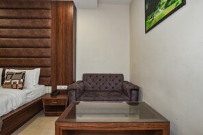 A hotel room with a bed, a wooden headboard, a grey sofa, a glass coffee table, and a painting on the wall. - Pineberry Hotels (Amritsar)