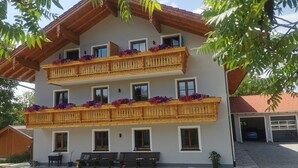 A multi-story building with wooden balconies adorned with flowers, a covered outdoor seating area, and a garage. - Nr. 1 Neubau Ferienwohnung mit 2 Sep. Schlafzimmern, Ost-balkon, 75qm (Prien am Chiemsee)