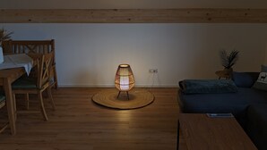 A cozy living room with a wooden floor, a round woven rug, a modern floor lamp, a wooden dining table with chairs, and a dark blue sofa with a decorative pillow. - Nr. 1 Neubau Ferienwohnung mit 2 Sep. Schlafzimmern, Ost-balkon, 75qm (Prien am Chiemsee)