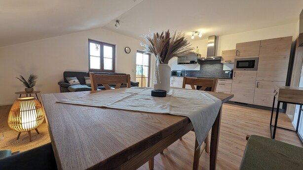 A modern kitchen with wooden cabinets, a dining area with a wooden table and chairs, and a cozy living room with a sofa and a lamp. - Nr. 1 Neubau Ferienwohnung mit 2 Sep. Schlafzimmern, Ost-balkon, 75qm (Prien am Chiemsee)