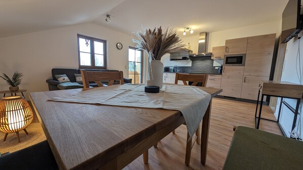 A modern kitchen with wooden cabinets, a dining table with a white tablecloth, and a vase with dried plants.