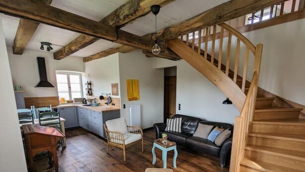 Un loft spacieux avec des poutres apparentes, une cuisine dotée d’un îlot central, un coin salon confortable avec un canapé et des fauteuils, et un escalier en bois.