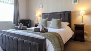 Una cama bien hecha con un cabecero tapizado en gris, dos mesitas de noche con lámparas, un sillón verde y una ventana con cortinas.