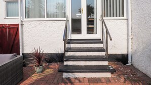 Una casa con fachada blanca, una escalera negra que lleva a una puerta de cristal y una pequeña planta en maceta en el suelo.