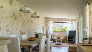 A dining area with stone walls, white furniture, a chandelier, and a view of greenery outside. - 3676 Villa Enò by Perle di Puglia (Noci)