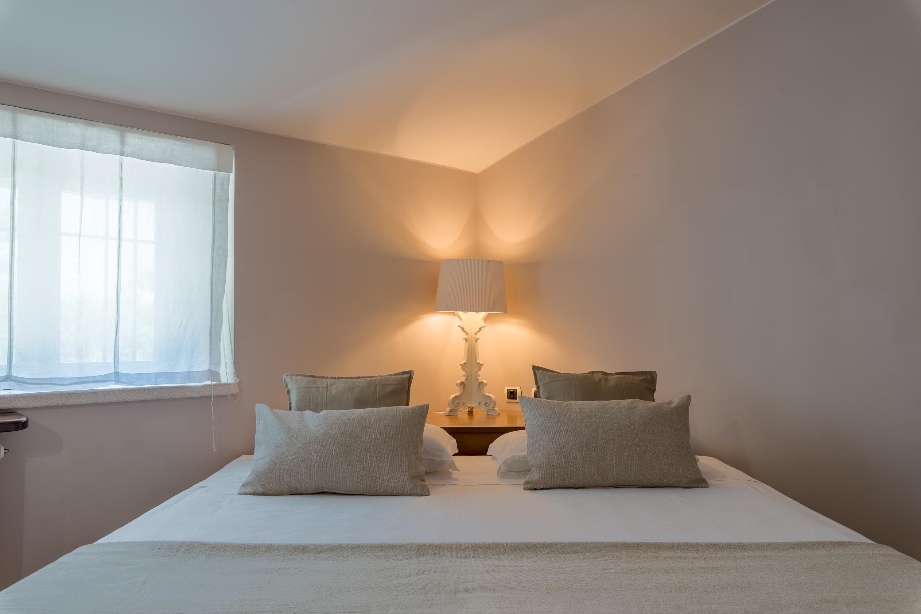 A neatly made bed with four pillows, a bedside table with a lamp, and a window with sheer curtains.