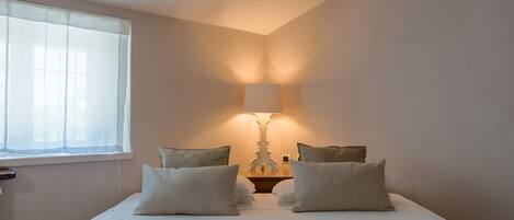 A neatly made bed with four pillows, a bedside table with a lamp, and a window with sheer curtains.