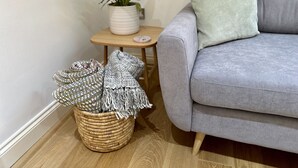 A grey sofa with a light green pillow, a small wooden side table with a potted plant and a white vase, and a wicker basket with blankets.