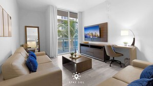 A modern living room with a sofa, coffee table, and a desk with a chair. There is a large window with a view of palm trees and a cityscape.