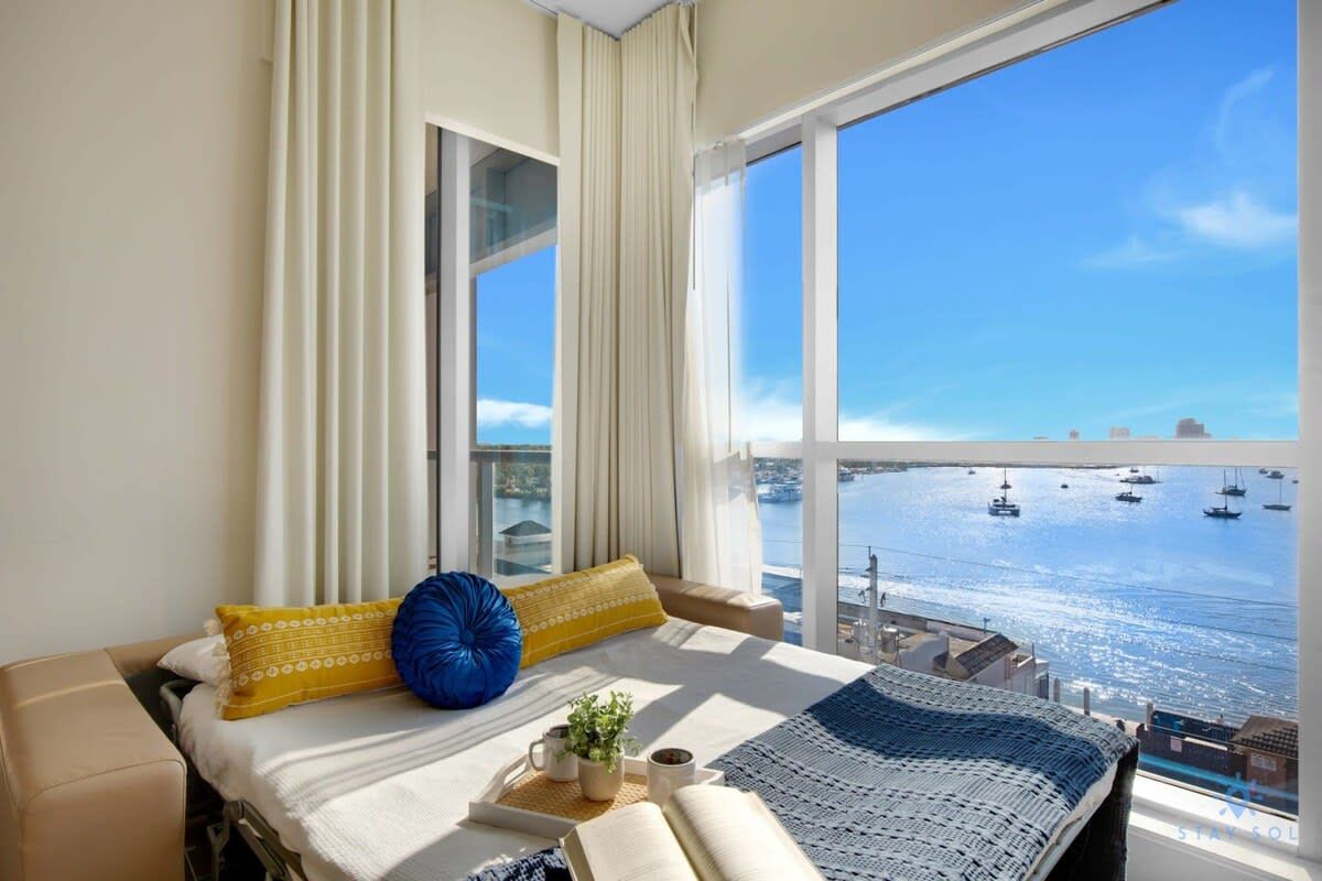 A hotel room with a large window, a bed with a blue and yellow pillow, a bedside table with a plant and a cup, and a view of boats on the water.