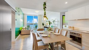 Un espace repas moderne avec une table et des chaises en bois, une cuisine dotée de meubles blancs et une vue sur la mer grâce à de grandes fenêtres.