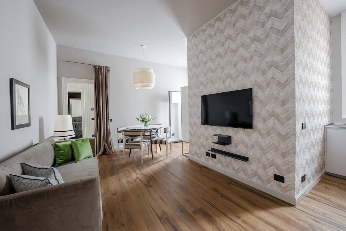 A modern living room with a flat-screen TV mounted on a chevron-patterned wall, a wooden floor, a sofa with green and grey cushions, a dining area with a table and chairs, and a large window with curtains.