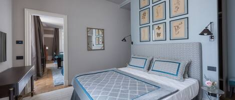 A bedroom with a large bed, a desk, and a chair. There are framed pictures on the wall and a lamp on the side table.