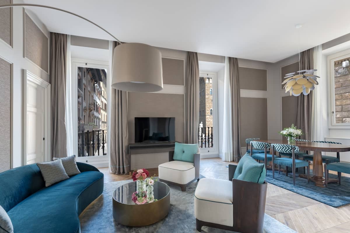 A modern living room with a sofa, armchairs, a coffee table with flowers, a TV, and a dining area with chairs and a table.