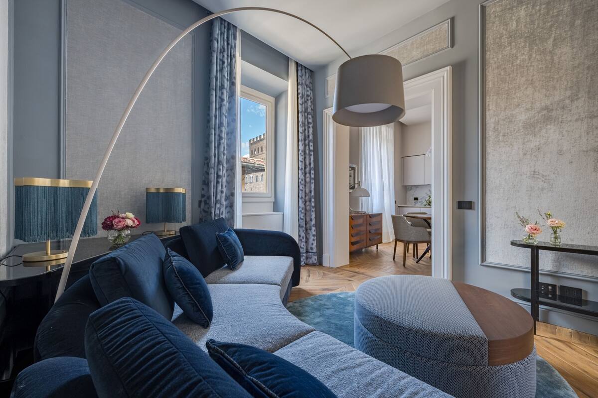 A modern living room with a sofa, a coffee table, and a lamp. There is a view of a cityscape through a window.