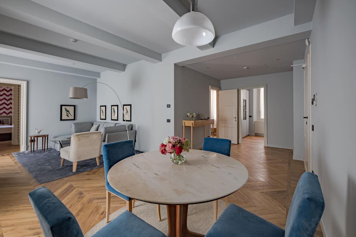 A spacious living room with a round table, blue chairs, and a vase of flowers. There is a sofa and a coffee table in the background.