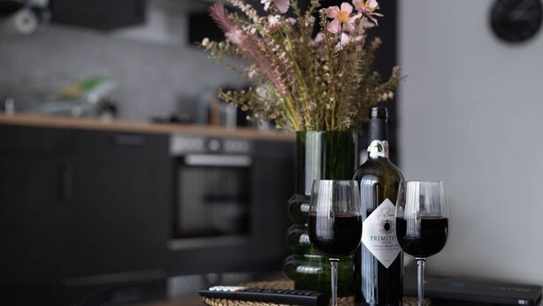 A kitchen with a vase of flowers, a wine bottle, and two glasses of red wine on a table. - Schicke Apartments in Osnabrück (Osnabrueck)