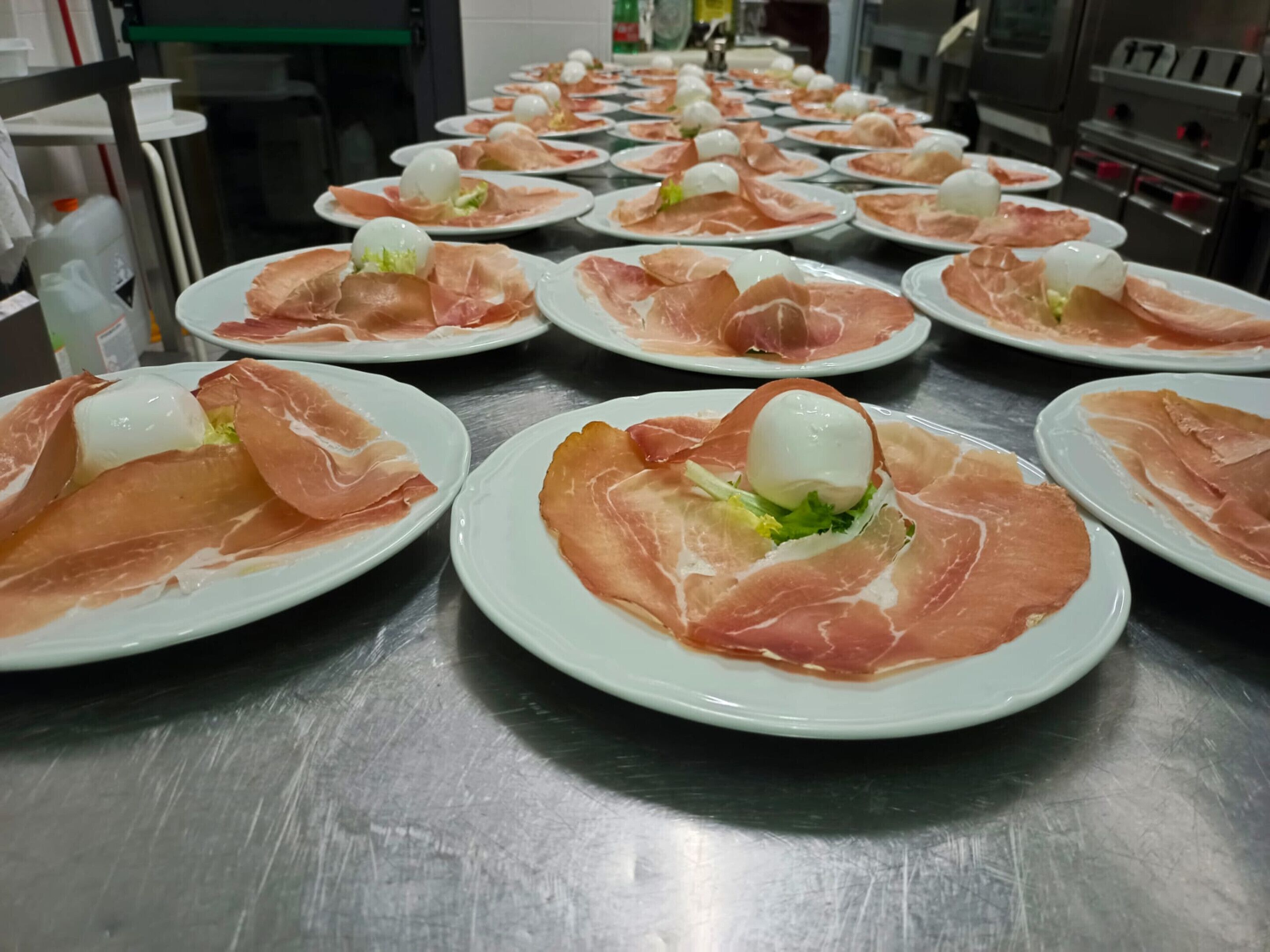 A kitchen with multiple plates of food, each topped with a slice of prosciutto and a ball of cheese, arranged on a stainless steel counter.
