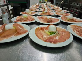 Una cocina con varios platos de comida, cada uno coronado con una loncha de jamón serrano y una bola de queso, dispuestos sobre una encimera de acero inoxidable.