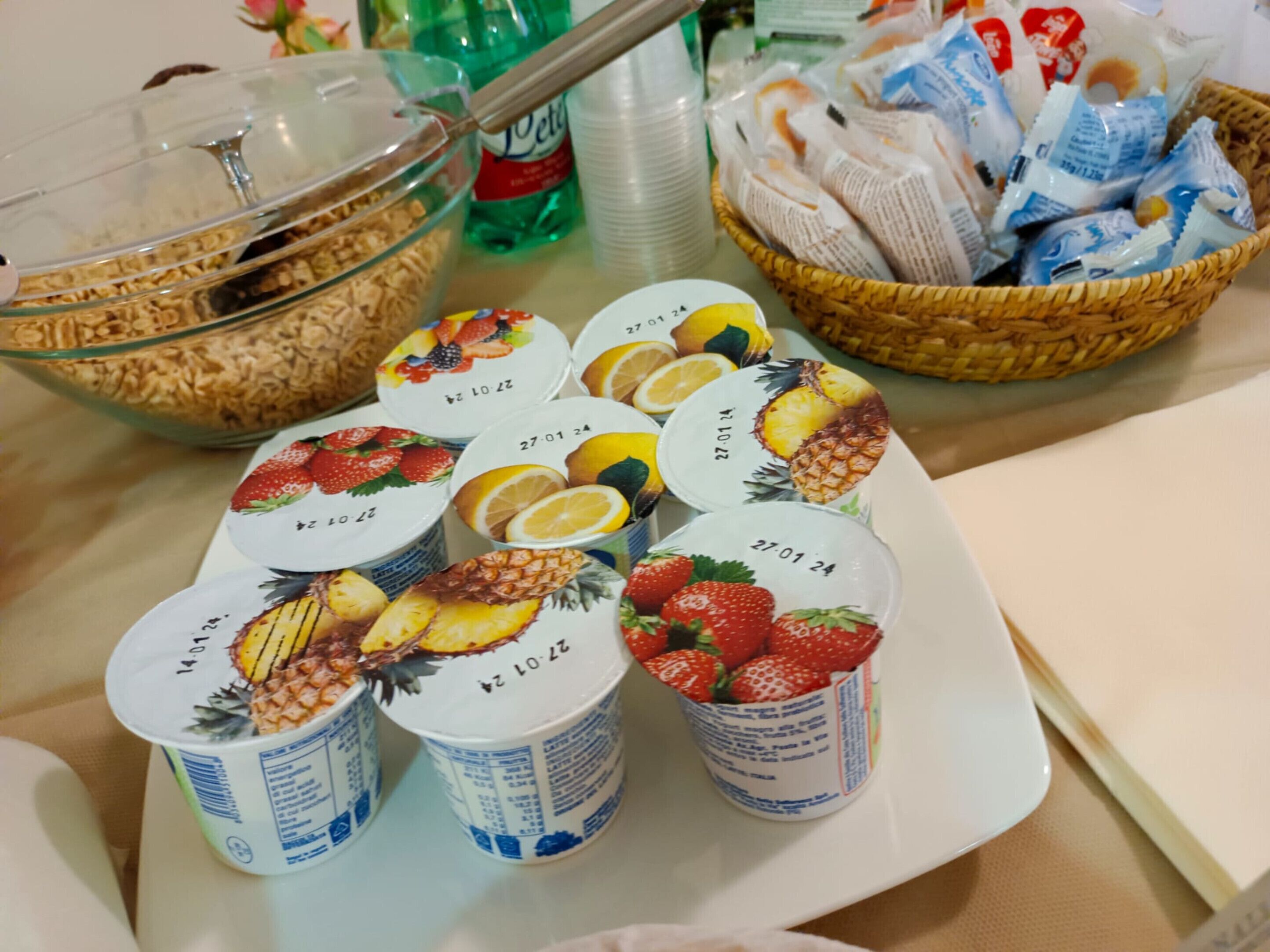 Une table sur laquelle sont disposés divers aliments, notamment un bol de céréales, un panier de pain et plusieurs tasses décorées de motifs de fruits.