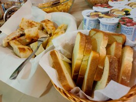 Una cesta con pan en rodajas y un tenedor sobre una mesa, junto a un tazón de cereal y recipientes de yogur.