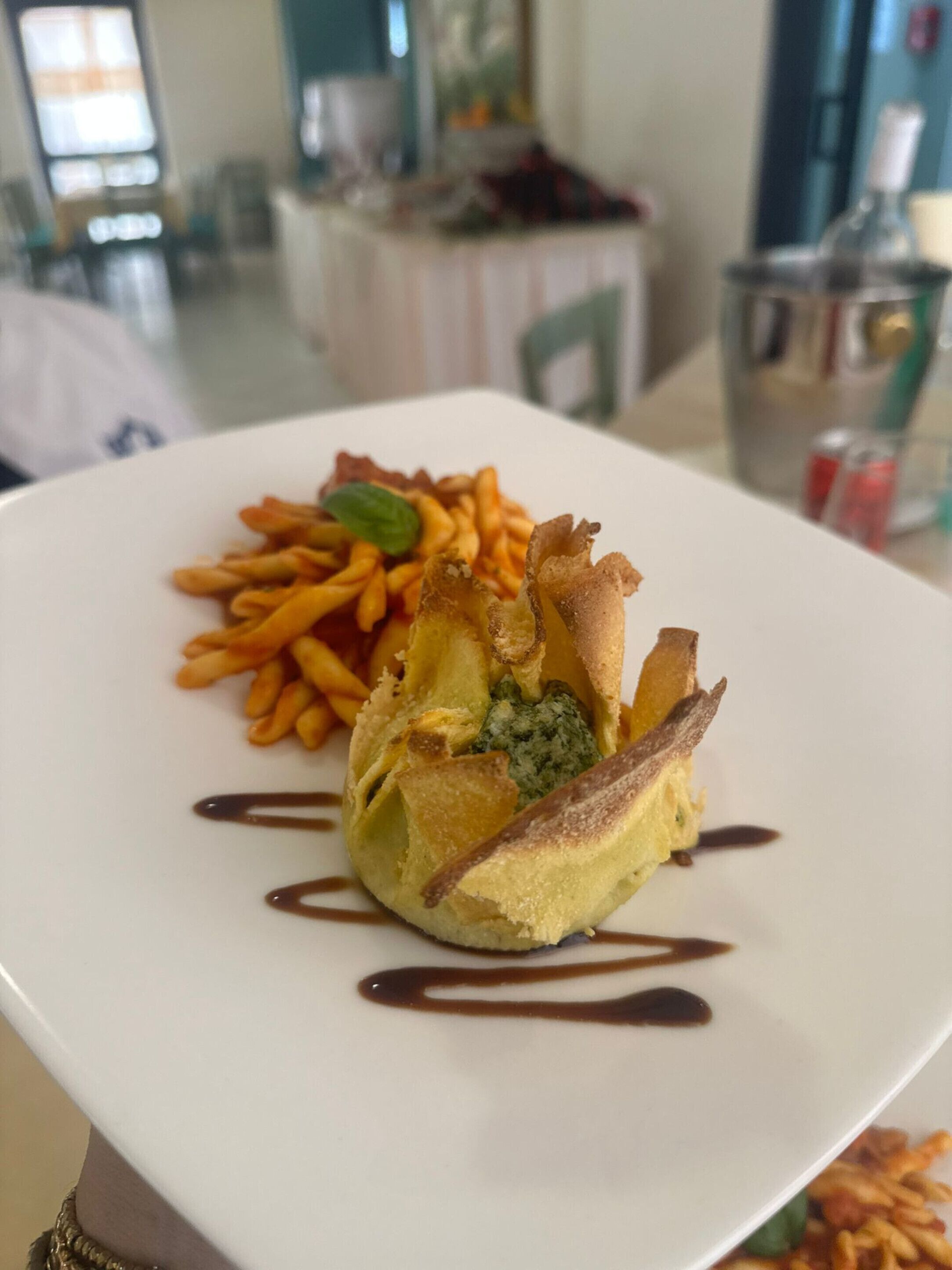 A white plate with a dessert consisting of a pastry, fries, and a green sauce, with a brown drizzle on a table in a restaurant.