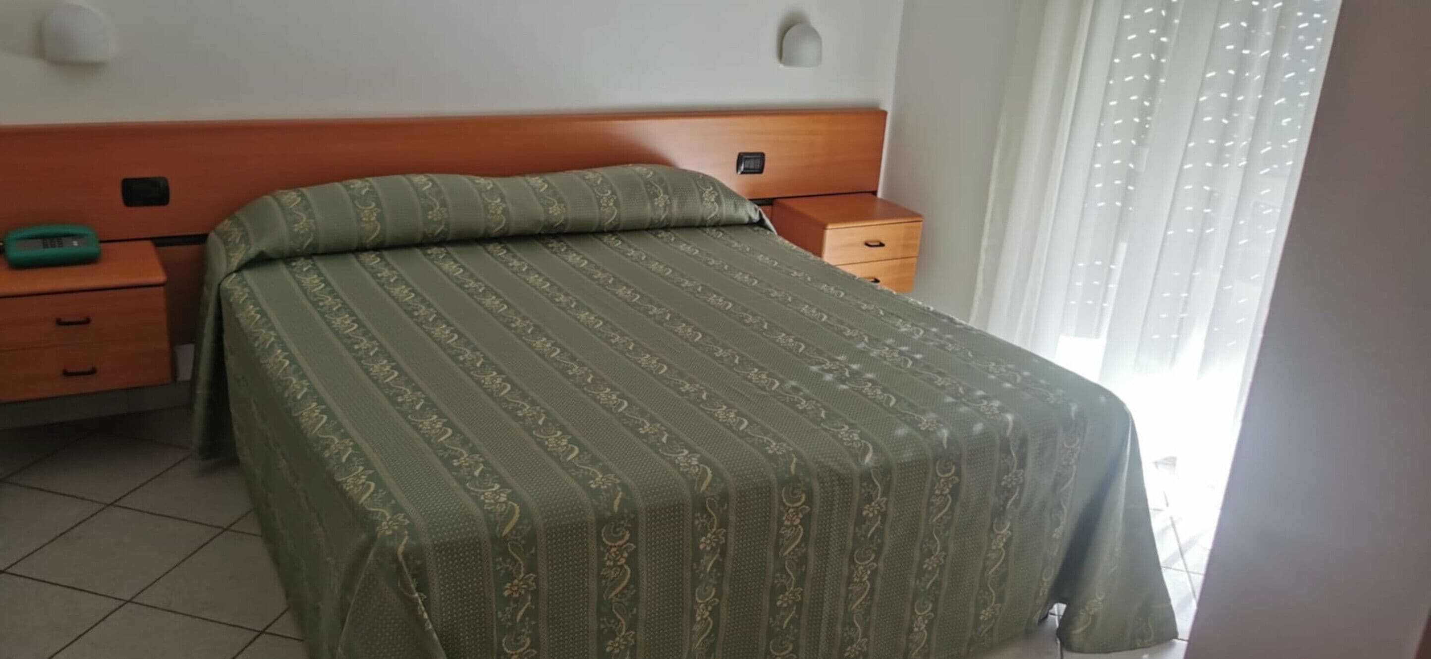 Una cama bien hecha con una colcha estampada, un cabecero de madera, una mesita de noche con un objeto verde y una ventana con cortinas.