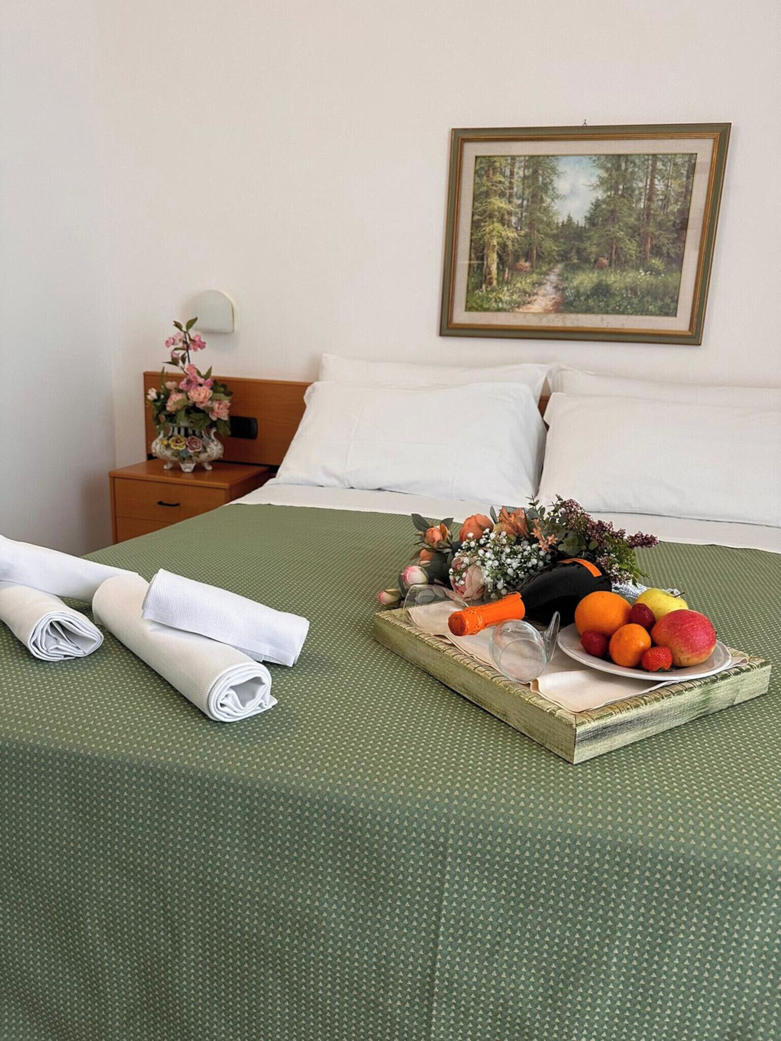 Cama bem arrumada com colcha verde, bandeja de frutas e toalhas dobradas em uma mesa de cabeceira de madeira. Um quadro com uma pintura de floresta na parede.