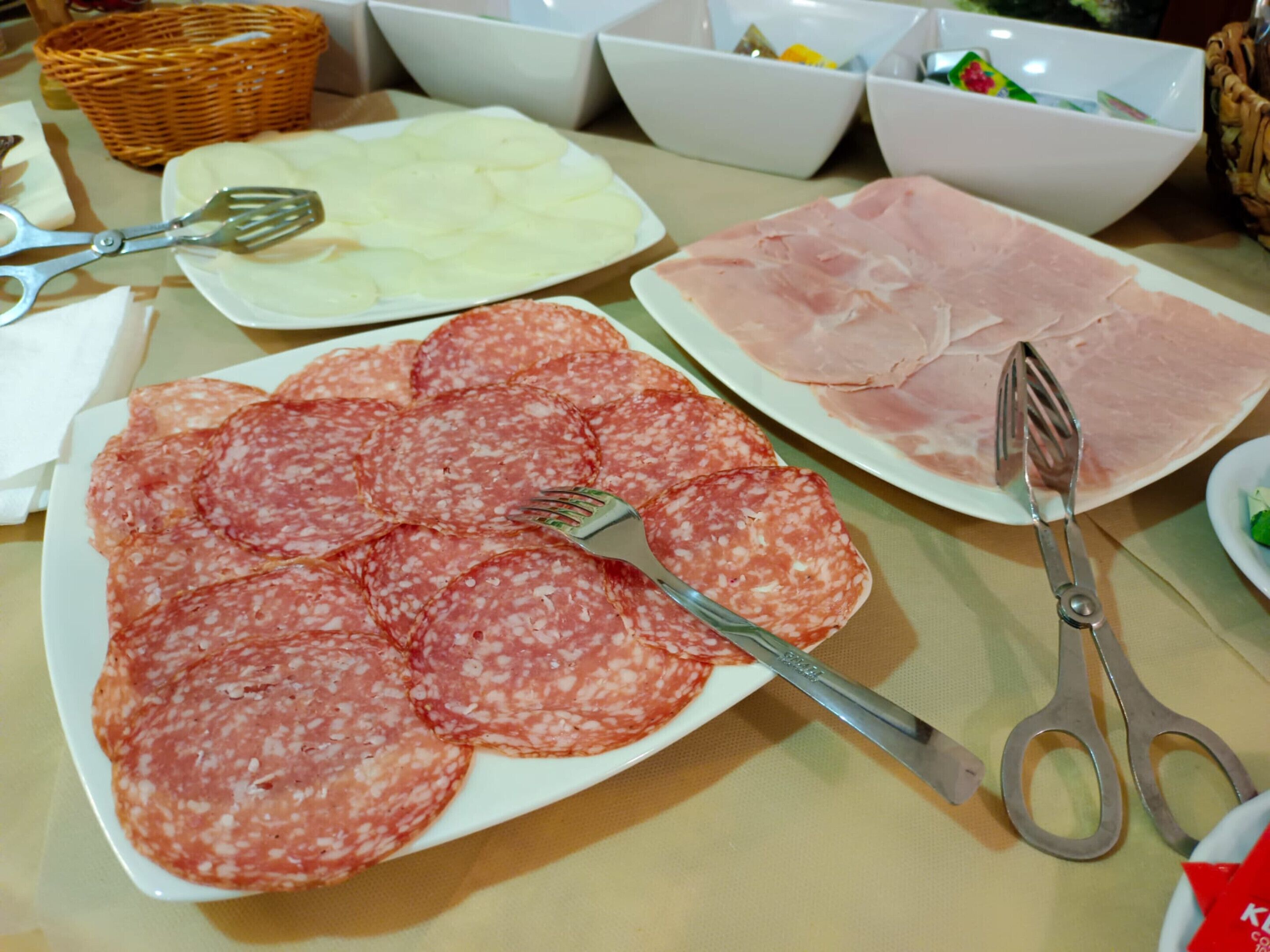 A table with plates of sliced salami and ham, a fork, and a pair of scissors.