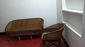 Twee rieten stoelen en een kleine bank in een hoek, met een rode vloer en witte wanden.