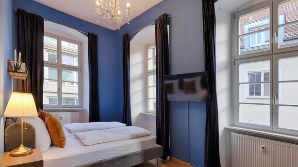 Un dormitorio con cama, mesita de noche, lámpara, araña y ventana con cortinas.