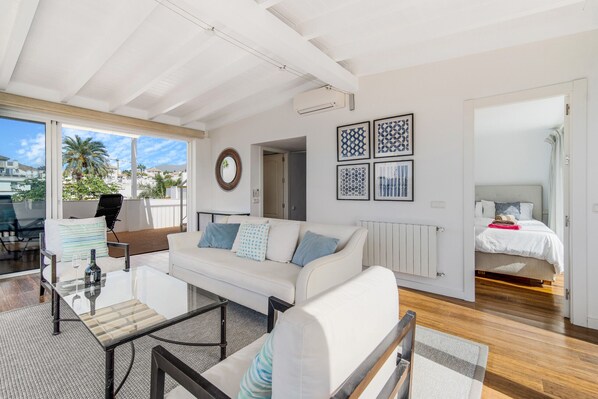 A modern living room with a white sofa, a glass coffee table, and a view of the outside. There is a bed in the adjacent room with a white headboard.
