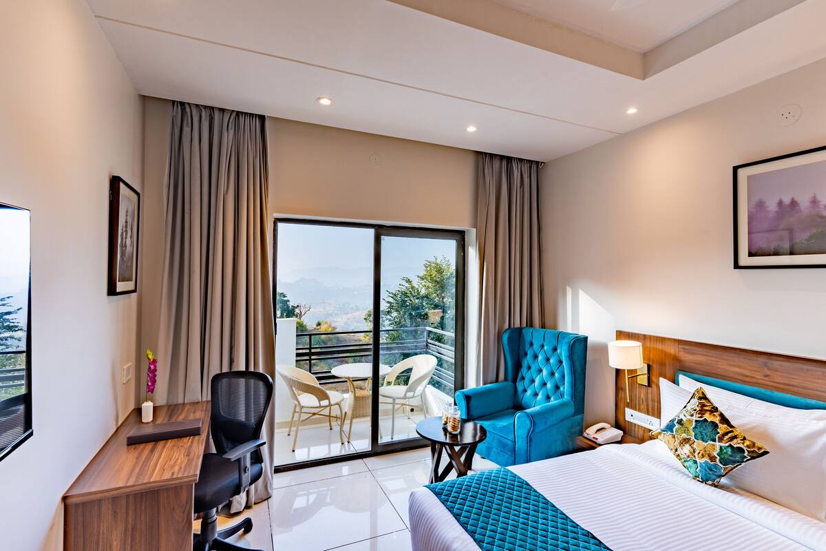 A hotel room with a bed, a desk, a chair, a small table, a lamp, and a view of the outdoors.