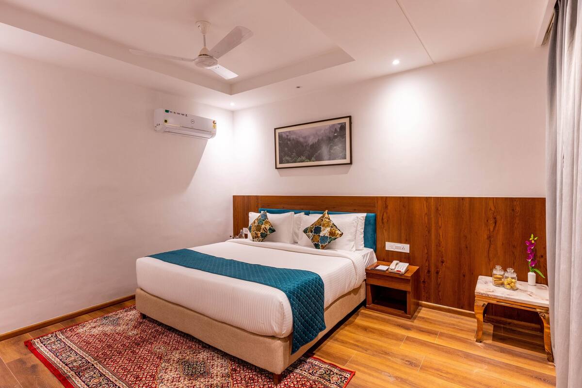 A hotel room with a bed, a nightstand, a wooden headboard, a ceiling fan, an air conditioning unit, a framed picture on the wall, and a small table with a vase and jars.
