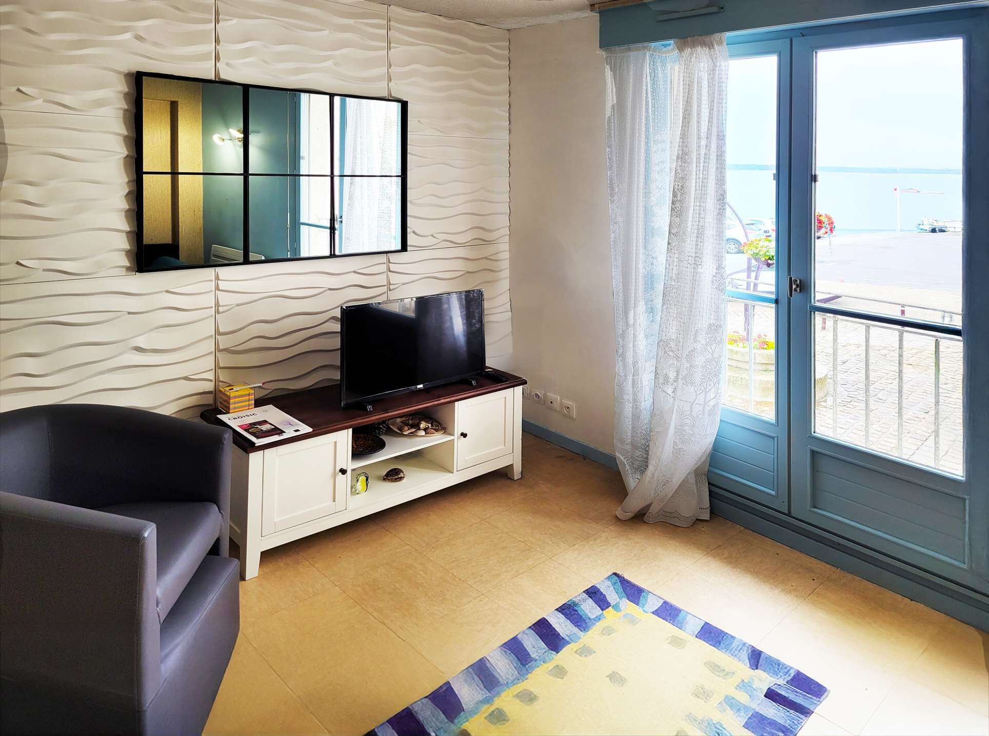A room with a television on a wooden stand, a white cabinet, a black chair, and a view of the sea through a glass door.