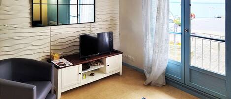A room with a television on a wooden stand, a white cabinet, a black chair, and a view of the sea through a glass door.