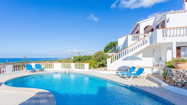 A pool area with a view of the sea, a white building with balconies, and blue lounge chairs. - Villa Lucas - Magnificent Sea Views (San Jaime Mediterráneo)