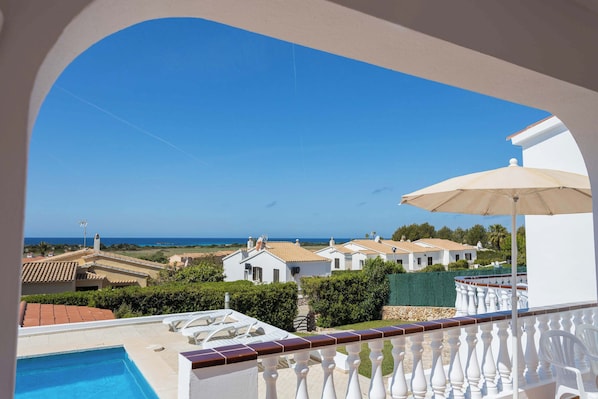 Une vue sur la piscine, avec un ciel dégagé, un parasol blanc et une rangée de bùtiments blancs en arriÚre-plan.