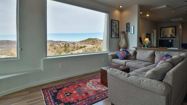 A living room with a large window offering a view of a desert landscape. There is a beige sofa with colorful pillows, a wooden coffee table, and a red patterned rug. - Moab Indian Hills - Secluded Breathtaking Views (Moab)