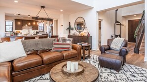 A spacious living area with a leather sofa, a coffee table, and a leather armchair. There is a kitchen in the background with modern appliances and pendant lighting.